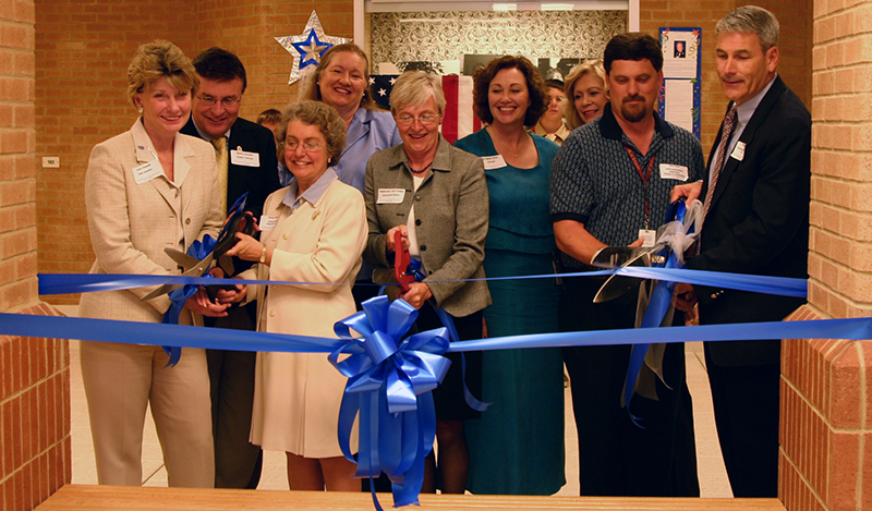 Photograph of the ribbon cutting ceremony during Colvin Run’s dedication. Local politicians, school staff, and FCPS officials cut a blue ribbon with two overly large scissors.
